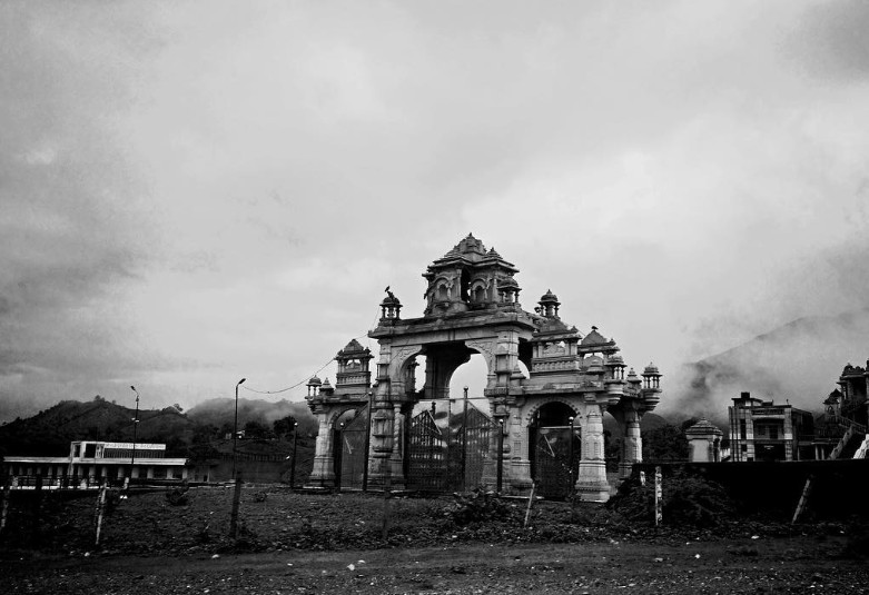 Ornate heritage gate with domes and carvings under monsoon clouds with hills behind